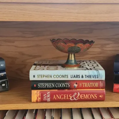 Sale Photo Thumbnail #272: Wooden bookshelf filled with stacks of popular fiction hardcovers, featuring authors like Steve Berry, Stephen Coonts, and Dan Brown, alongside decorative tabletop items.