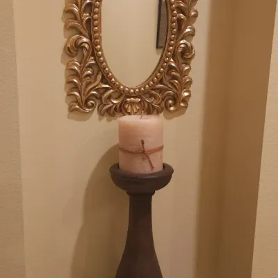 Sale Photo Thumbnail #131: Ornate gold-finished wall mirror with scrollwork frame displayed above a tall wooden candlestick pedestal and pillar candle set in a wall niche.