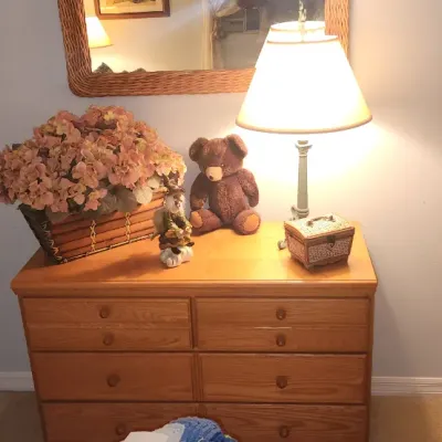 Sale Photo Thumbnail #177: Light oak wooden dresser featuring six drawers, topped with decor including a large faux flower arrangement, a tabletop lamp, and a teddy bear.