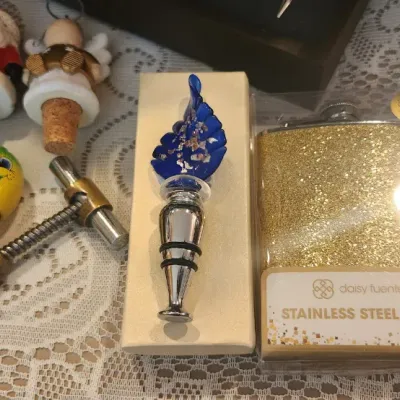 Sale Photo Thumbnail #146: Collection of barware accessories including decorative wine stoppers, a vintage brass corkscrew, and a boxed Daisy Fuentes gold glitter stainless steel flask.