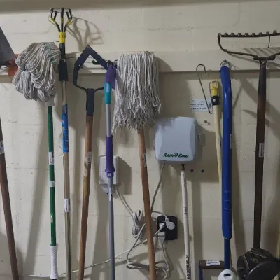 Sale Photo Thumbnail #350: Assorted long-handled yard and cleaning tools including shovels, mops, rakes, and a pole pruner hanging on a wall alongside a Rain Bird irrigation controller.