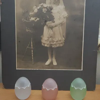 Sale Photo Thumbnail #302: Early 20th-century sepia studio portrait of a girl in a frilly dress holding flowers, displayed behind three cracked egg-shaped frosted glass containers and a hand fan on a lace runner.