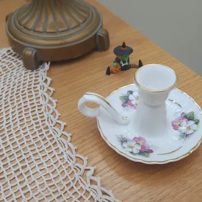Sale Photo Thumbnail #304: Vintage white porcelain chamberstick candlestick holder with pink floral motifs and gold rim details, sitting on a wooden surface beside a crocheted doily.