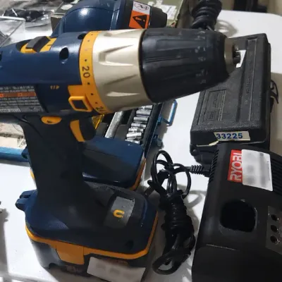 Sale Photo Thumbnail #376: Ryobi 18V cordless drill with battery pack and charger, shown alongside various household power tools and accessories on a table.
