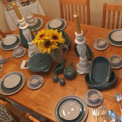 Sale Photo Thumbnail #9: Oak dining table set with a large service of blue and grey ceramic tableware, accompanied by carved candlestick holders and silver-tone serving utensils.