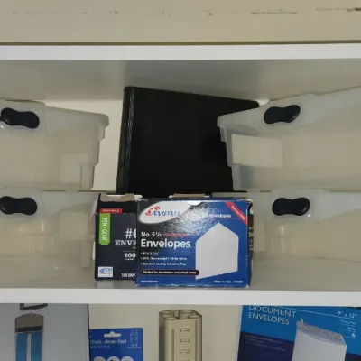 Sale Photo Thumbnail #286: Assortment of clear plastic storage totes with black handles, photo album, and boxes of mailing envelopes neatly arranged on white shelving.