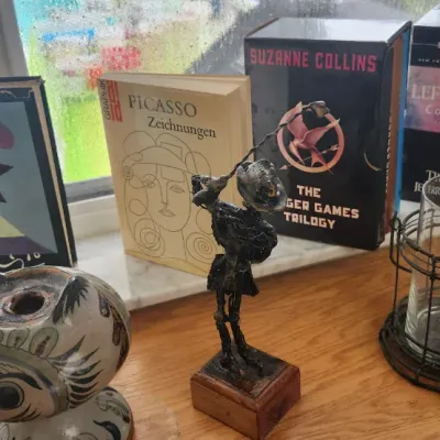 Sale Photo Thumbnail #275: Collection of books and decorative home accents displayed on a windowsill, including a Picasso art book, box sets of The Hunger Games and Left Behind, a small metal Don Quixote figurine, and a hand-painted ceramic owl vessel.