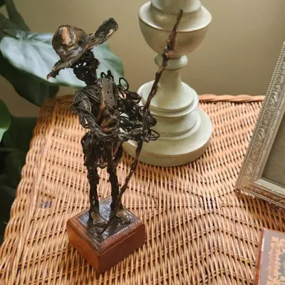 Sale Photo Thumbnail #265: Welded wire metal sculpture of Don Quixote on a wooden base, displayed alongside an ornate picture frame on a wicker surface.