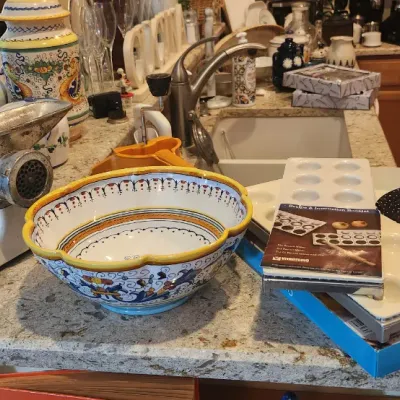Sale Photo Thumbnail #213: Kitchenware collection including a hand-crank meat grinder, an Italian hand-painted ceramic bowl, a ravioli maker mold with instruction booklet, and a black and white ceramic mug.