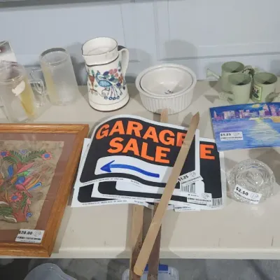 Sale Photo Thumbnail #341: Assorted household items displayed on a folding table, including framed artwork, a hand-painted ceramic pitcher, garage sale signs, glassware, and barbecue utensils.