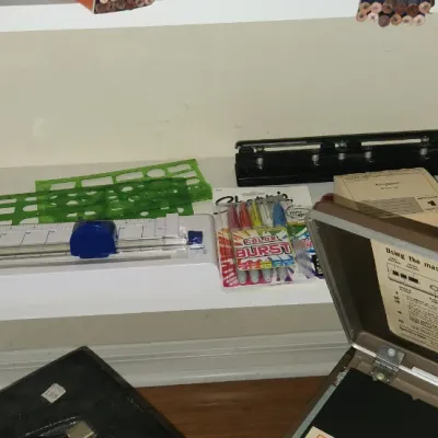 Sale Photo Thumbnail #284: Collection of office and crafting supplies including a paper trimmer, hole punch, green stencils, and Sharpie markers, displayed on shelving.