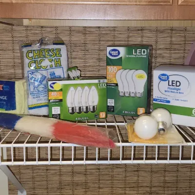 Sale Photo Thumbnail #248: Assortment of household cleaning supplies, lightbulbs, and miscellaneous items arranged on a wire shelf. Includes packaged Great Value LED bulbs, a Spontex sponge, cheesecloth, and a duster.