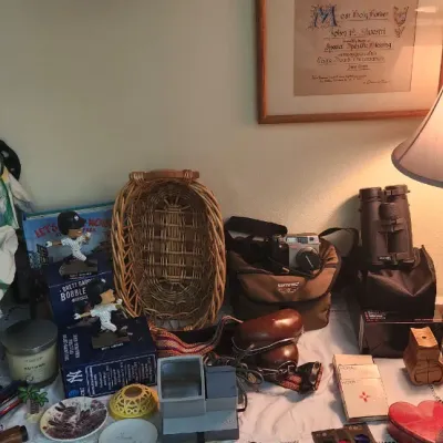Sale Photo Thumbnail #295: Collection of tabletop decor and collectibles including a jester doll, baseball bobbleheads, woven basket, point-and-shoot cameras, binoculars, and a lighted table lamp with ribbon accent.