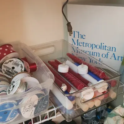 Sale Photo Thumbnail #293: An assortment of crafting supplies, including clear bins of spooled ribbons and taper candles, alongside a Metropolitan Museum of Art box and a cloth doll.