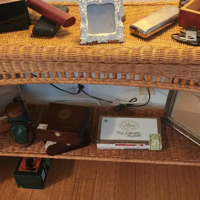 Sale Photo Thumbnail #255: Woven wicker console table with a lower shelf, displaying various wooden cigar boxes, picture frames, and small smoking accessories.