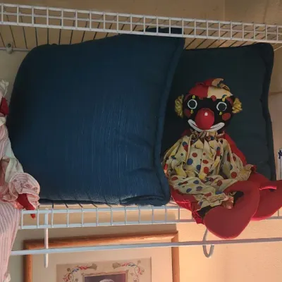 Sale Photo Thumbnail #290: Two vintage fabric clown dolls sitting on a wire shelf alongside decorative throw pillows and a boxed New York Yankees item.