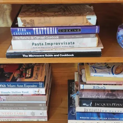 Sale Photo Thumbnail #211: Selection of culinary and Italian cookbooks displayed on a wooden shelf alongside a blue glass bottle and a decorative patterned tin.