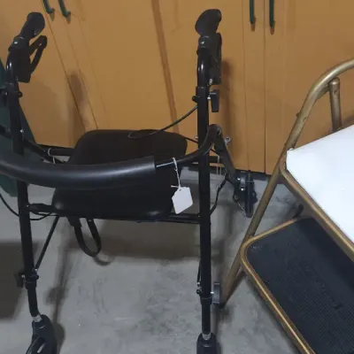 Sale Photo Thumbnail #361: Black rolling walker with seat and handbrakes alongside a vintage gold-toned metal step stool chair with a cushioned top.