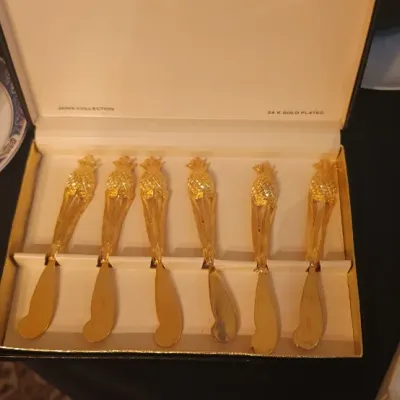 Sale Photo Thumbnail #15: Boxed set of six 24K gold-plated pineapple-motif butter spreaders by Janis Collection, displayed alongside porcelain teacups and saucers.