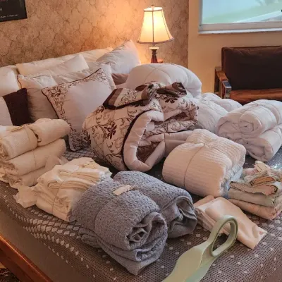 Sale Photo Thumbnail #104: Large collection of bed linens, comforters, and throw pillows displayed on a bed frame, along with traditional bedside lamps and a wicker loveseat.