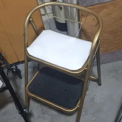 Sale Photo Thumbnail #360: Vintage gold-toned metal step stool chair with a cushioned white seat and black rubber lower step.