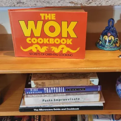 Sale Photo Thumbnail #210: Collection of cookbooks including 'The Wok Cookbook', displayed on wooden shelving alongside decorative ceramic bells, figurines, and a round patterned tin.