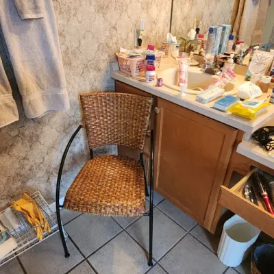 Sale Photo Thumbnail #115: Bathroom vanity area featuring a woven rattan armchair, a wire storage rack with towels, and various toiletries and hair styling tools displayed on the counter and in open drawers.