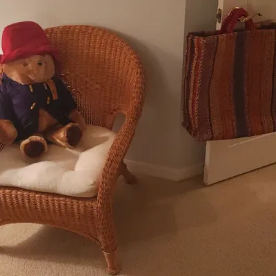 Sale Photo Thumbnail #174: Natural wicker armchair with a cream seat cushion, featuring a Paddington Bear plush toy and a striped woven tote bag hanging nearby.