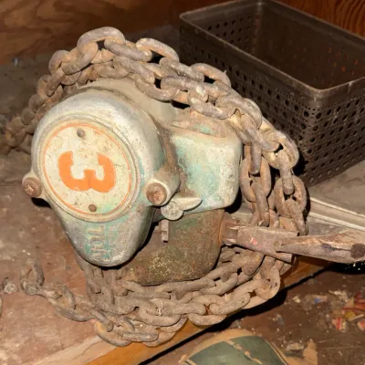 Sale Photo Thumbnail #43: Vintage heavy-duty 3-ton chain hoist with heavy steel chain wrapped around the body, showing signs of wear and surface rust.