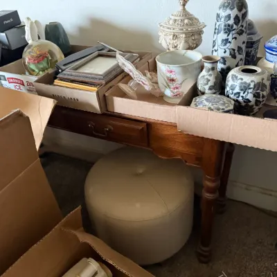 Sale Photo Thumbnail #42: Collection of home decor items displayed in cardboard flats atop a wooden side table, including blue and white ceramic vases, picture frames, and glassware, accompanied by a beige tufted ottoman.