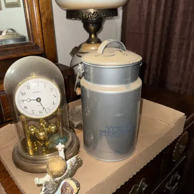 Sale Photo Thumbnail #11: Vintage Howard Miller anniversary dome clock alongside a decorative silver-tone metal milk can container and a small angel figurine.