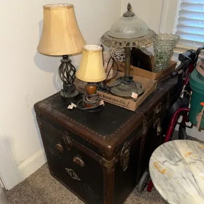 Sale Photo Thumbnail #8: Vintage dark leather and metal-bound steamer trunk supporting a assortment of table lamps, including a brass lamp with a frosted glass shade.