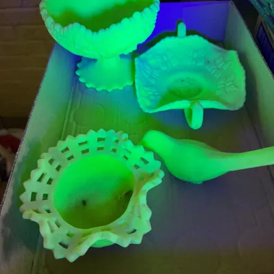 Sale Photo Thumbnail #32: Collection of four vintage fluorescent uranium glass pieces, including a bird figurine, a open lace ruffle edge bowl, and decorative candy dishes, glowing brightly under blacklight.