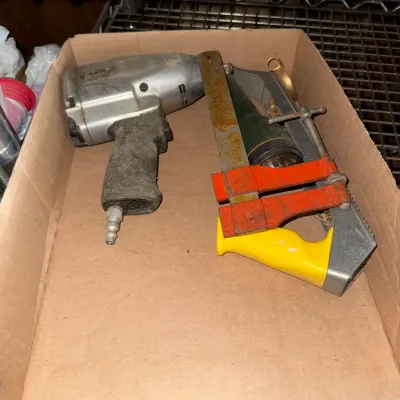 Sale Photo Thumbnail #46: Cardboard tray containing a pneumatic impact wrench and a yellow-handled hacksaw alongside an orange corner clamp.