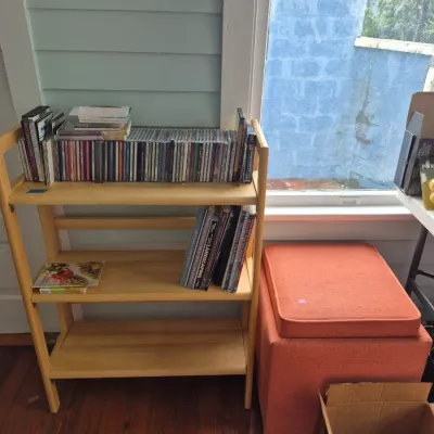 Sale Photo Thumbnail #31: Light wood three-tier bookshelf loaded with CDs and books, displayed next to an orange fabric storage ottoman.