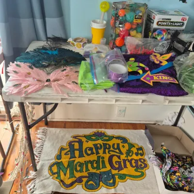 Sale Photo Thumbnail #82: Collection of Mardi Gras decorations and accessories, including feather masks, sequined decor, colorful beads, light displays, and a 'Happy Mardi Gras' fringe mat.