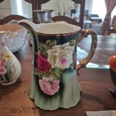 Sale Photo Thumbnail #2: Hand-painted porcelain pitcher featuring large pink and white rose motifs with gilt trim and a scroll handle, circa early 20th century.