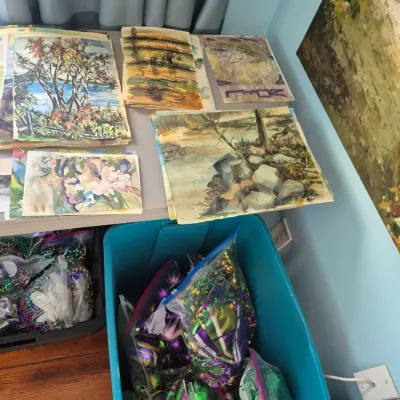 Sale Photo Thumbnail #74: A table arranged with original unframed watercolor and mixed-media paintings, displayed above plastic bins filled with Mardi Gras beads and festive decorations.