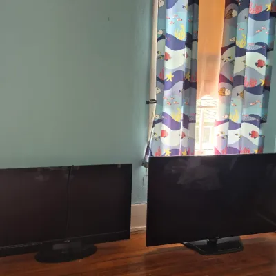 Sale Photo Thumbnail #95: Two black flat-screen computer monitors resting on a wooden floor in front of a window with ocean-themed curtains.