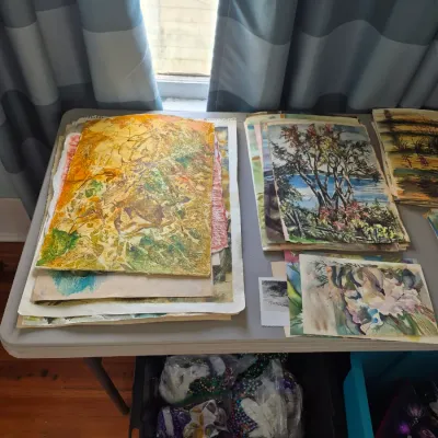 Sale Photo Thumbnail #79: Assortment of original unframed art prints and paintings arranged on a folding table, featuring landscapes and floral motifs.
