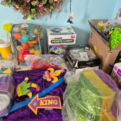 Sale Photo Thumbnail #83: Assortment of Mardi Gras party supplies and decorations, including a tinsel wreath, sequined fabric, glitter top hats, string lights, and novelty signs.