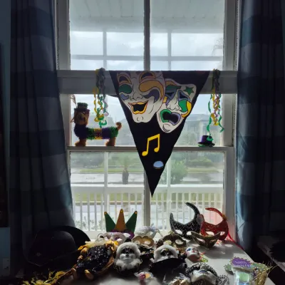 Sale Photo Thumbnail #77: Collection of Mardi Gras decorations including ornate masquerade masks, jester figurines, bead necklaces, and a comedy and tragedy banner arranged on a folding table in front of a window.