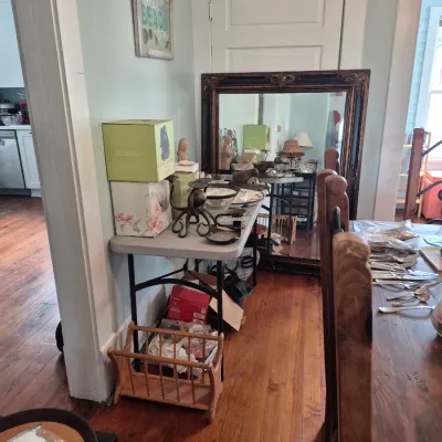 Sale Photo Thumbnail #44: Large dark ornate framed mirror sitting leaning against the wall alongside folding tables filled with decorative items, glassware, and tableware.