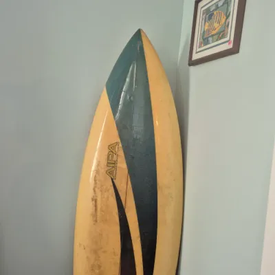 Sale Photo Thumbnail #52: Vintage Aipa sting surfboard featuring a yellow, dark green, and red graphic design, displayed leaning in a corner next to a framed fish art print.