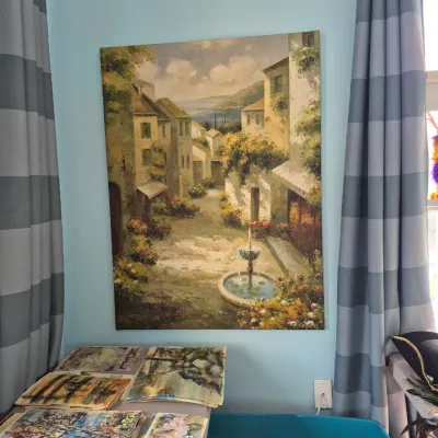 Sale Photo Thumbnail #76: Large unframed canvas print or painting of a European village scene with a central fountain, mounted on a light blue wall alongside stacked art prints.