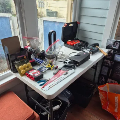 Sale Photo Thumbnail #29: Assorted power tools, hand tools, and hardware supplies arranged on a white folding table, including a jigsaw in a case, a cordless drill, a handsaw, pliers, and tape.