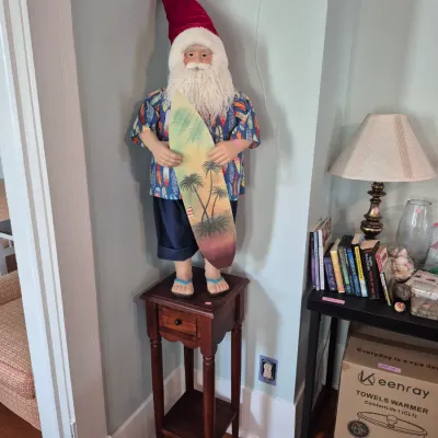Sale Photo Thumbnail #41: Lifesize surfing Santa figure dressed in a Hawaiian shirt and holding a palm tree surfboard, standing atop a dark wood plant stand.