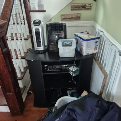 Sale Photo Thumbnail #54: Black wooden corner media console loaded with portable space heaters, including Sylvania and Lasko models, along with boxed electronics and miscellaneous household goods.