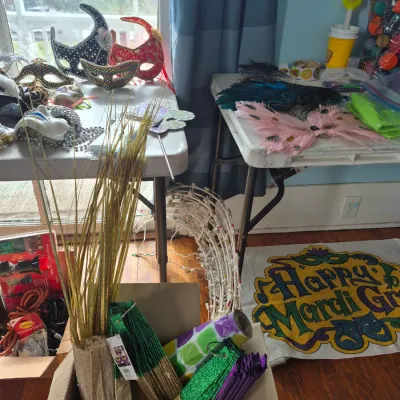 Sale Photo Thumbnail #81: Assortment of Mardi Gras party supplies and decor, including ornate masquerade masks, feathered wings, paper fans, decorative gold sprays, and a printed welcome mat.