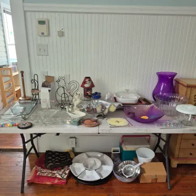 Sale Photo Thumbnail #15: Folding utility table filled with various home goods including purple art glass pieces, serving trays, Pyrex baking dishes, and kitchen strainers.